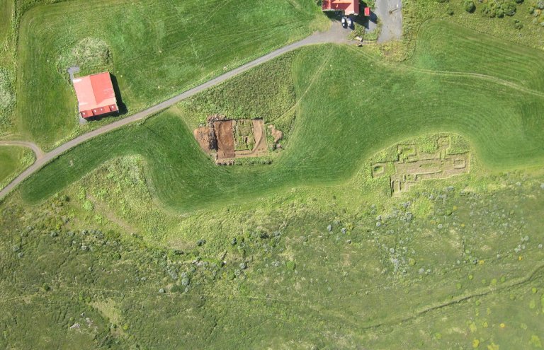 Loftmynd tekin með þyrlu, austan við túnið, sem sýnir afstöðu kirkjugarðsins gagnvart núverandi bæjarhúsum á Hofstöðum og rúst víkingaaldarskálans í ágúst árið 2013. ©Fornleifastofnun Íslands ses.